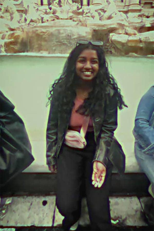 Reueline Arulanandam, a female student smiles at the camera while sitting on the edge of an ornate fountain. The photo has been heavily filtered to look like a vintage film photo.